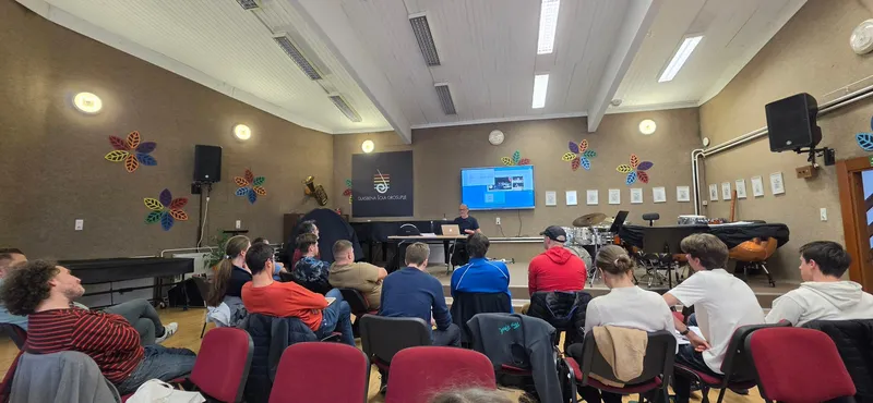 Širok pogled na dvorano Glasbene šole Grosuplje od zadaj: udeleženci sedijo v vrstah s hrbtom proti objektivu, v ozadju Lojze Krajnčan za mizo s prenosnikom pod zaslonom.