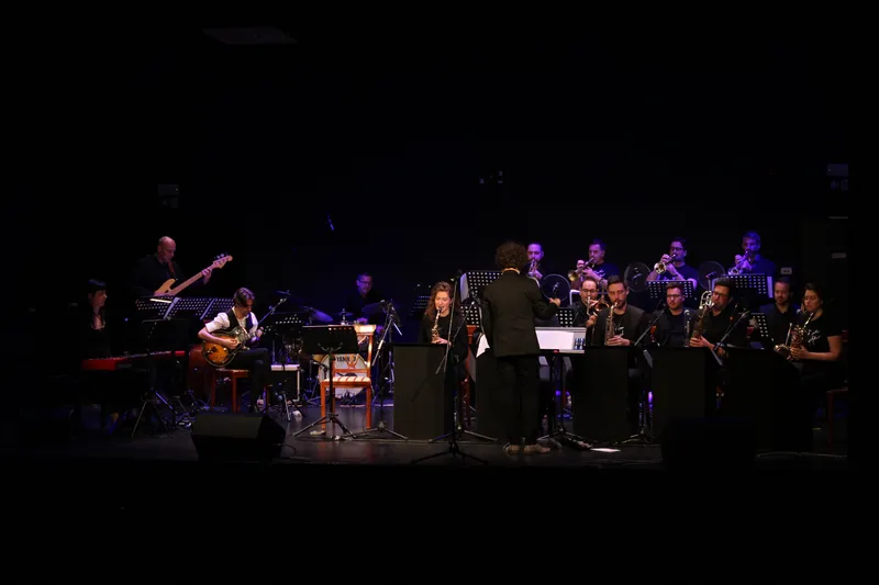 Big band v celoti na zatemnjenem odru: ritem sekcija s klaviaturami, kontrabasom, kitaro in bobni na levi, pihalne sekcije za pulti na desni, dirigent pred njimi.