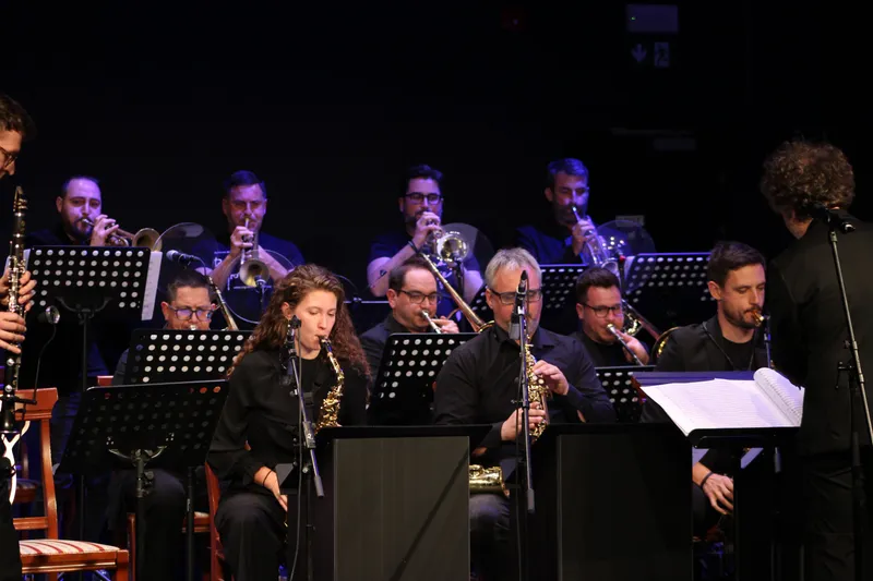 Orkester med igranjem: saksofoni v prvi vrsti pri mikrofonih, trobente in trombon v drugi, ob desnem robu kadra dirigent nad partituro.