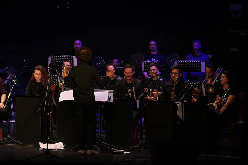 Sproščen trenutek med skladbama: dirigent z dvignjeno roko pri pultu, nasmejani člani orkestra z inštrumenti v rokah.