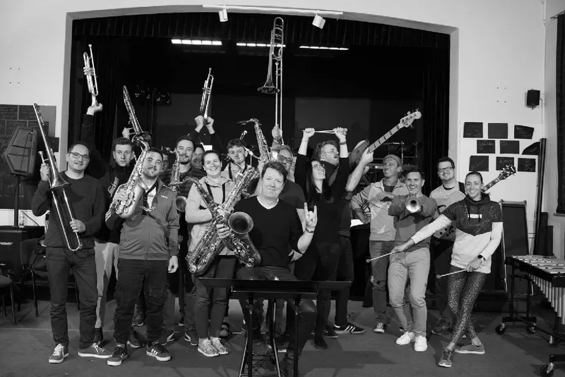 Črno-bela skupinska fotografija po vaji: orkester in gostujoči glasbeniki stojijo na odru z dvignjenimi inštrumenti — trombonami, trobentami in saksofoni; na desni vibrafon, na sredini sedi dirigent za pultom.
